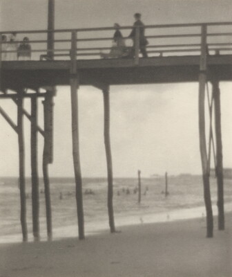 A black-and-white photograph looking up at people walking on a tall pier that sits over a beach.