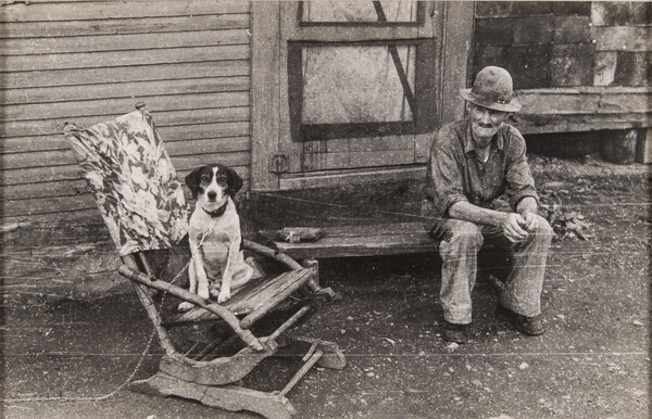 A black-and-white photograph of an elderly man in ragged clothing sitting on a wood stoop as a dog sits in a wood rocker next to him.