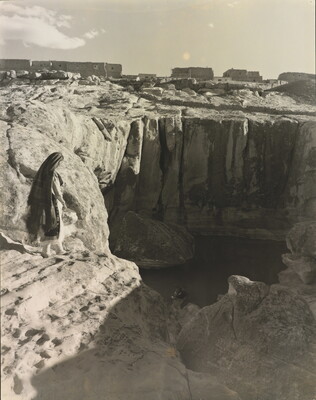 A black-and-white photograph of a woman with a long headscarf on a cliff, overlooking a water hole at the bottom.