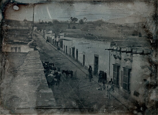 A black-and-white photograph, slightly obscured by surface marks, of a street lined with buildings, people and horses.