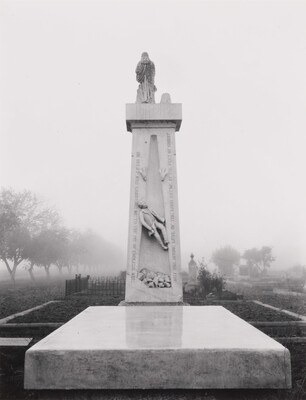 A black-and-white photograph of a tall, vertical stone monument rising behind a flat stone slab in a cemetery.