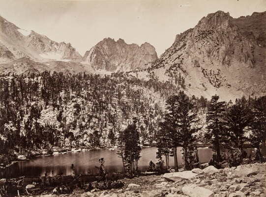 A black-and-white photograph of a lake in a mountain valley surrounded by pine trees and mountains dusted in snow.