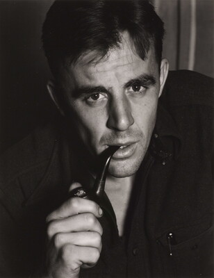 A black-and-white portrait photograph of a White man in a dark shirt leaning forward as he smokes a pipe.