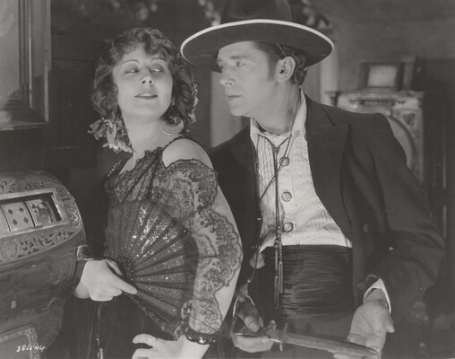 A black-and-white photograph of a White woman wearing a 1920s-style dress holding an open fan against her shoulder as the White man behind her holds a dagger.