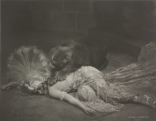 A black-and-white photograph of an elaborately dressed White woman lying on the ground with a snarling lion lying over her back.