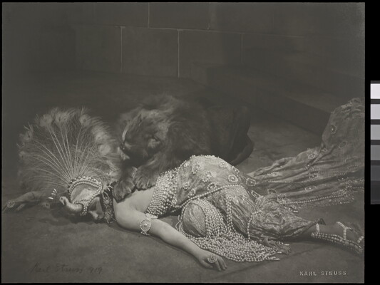 A black-and-white photograph of an elaborately dressed woman lying on the ground with a person in a lion costume lying over her back.