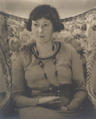 A black-and-white photograph a White woman with short dark hair wearing a long necklace and a long-sleeve top sitting in a floral, wingback chair.