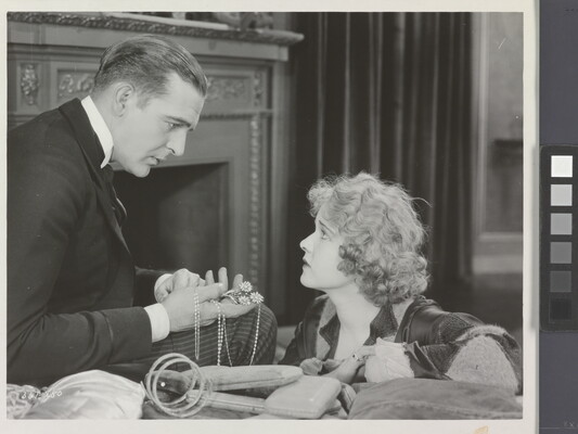 A black-and-white photograph of a White man presenting a White woman with jewelry as she gazes up at him looking worried.