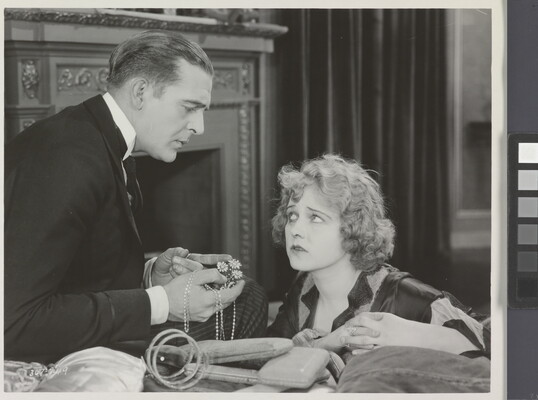 A black-and-white photograph of a White man presenting a White woman with jewelry as she gazes up at him looking worried.