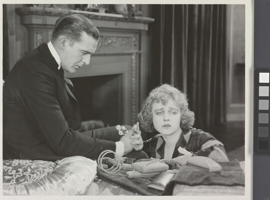 A black-and-white photograph of a White man presenting a White woman with jewelry as she sadly gazes into the distance.