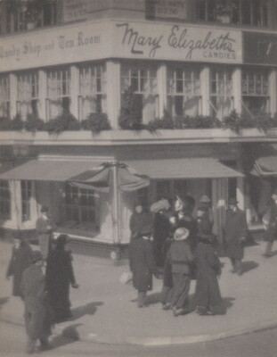 A black-and-white photograph of people walking along a street corner in front of a multi-story building with awnings at street level.