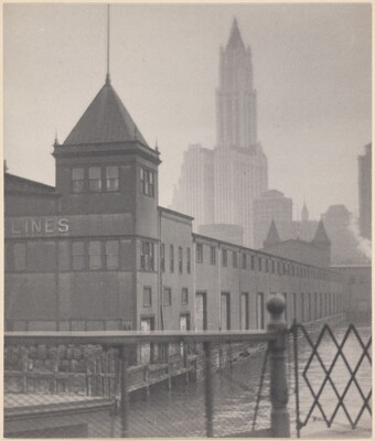 A black-and-white photograph of a low building with a tower on a canal, a gate in the foreground, and skyscrapers in the background.