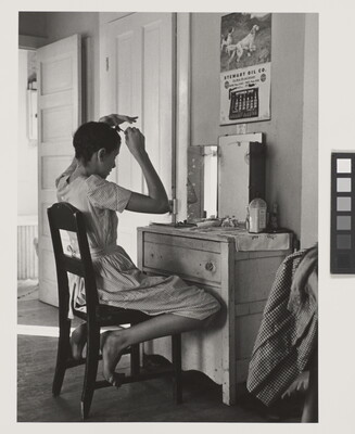 A black-and-white photograph of a White teenage girl seated in front of a mirror on top of a dresser fixing her hair.