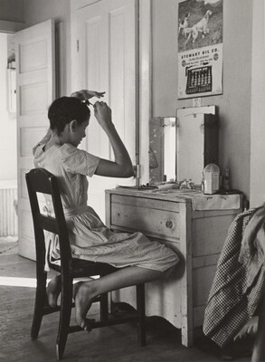 A black-and-white photograph of a White teenage girl seated in front of a mirror on top of a dresser fixing her hair.