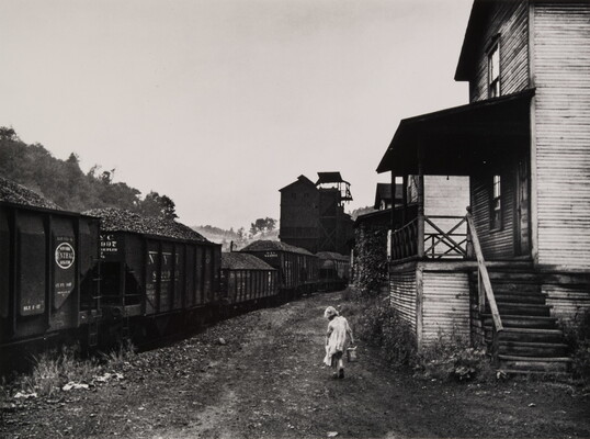 A black-and-white photograph of a young girl carrying a pail between a train and some clapboard buildings.