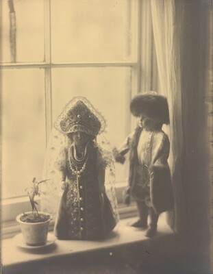 A sepia-toned photograph of two light-colored dolls in Russian imperial clothing standing next to a flower pot on a window sill.