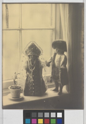 A sepia-toned photograph of two light-colored dolls in Russian imperial clothing standing next to a flower pot on a window sill.