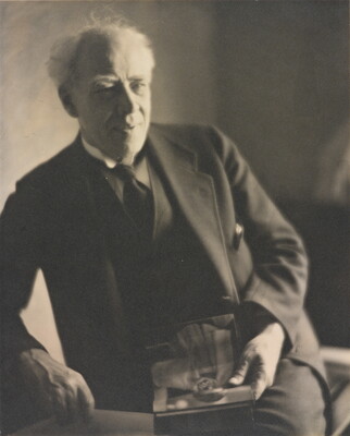 A black-and-white photograph of an older White man with white hair, wearing a dark suit and tie, leaning on one arm while holding an object in his hand.