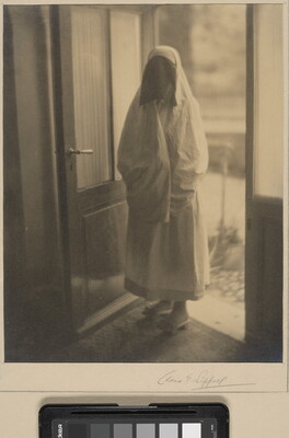 A black-and-white photograph of a woman in a doorway covered from head to toe in a white niqab with dark netting over her face.