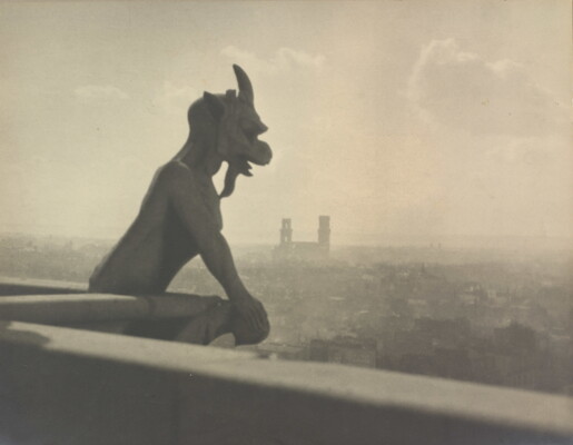A black-and-white photograph of a gargoyle with horns and a long beard on the rooftop of a building overlooking a hazy city.