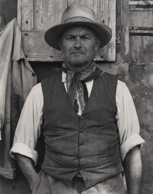 A black-and-white portrait photograph of a medium-skinned man from the waist up wearing a hat, bandanna tied around his neck, vest, and a shirt rolled to his elbows.