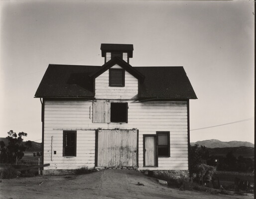 A black-and-white photograph of a white, two-story clapboard barn set against rolling hills with a small cat reclining in the dirt path.