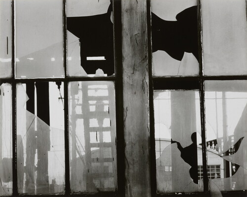 A black-and-white photograph of broken window panes in what looks like a factory building.