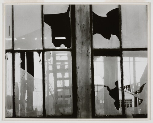 A black-and-white photograph of broken window panes in what looks like a factory building.