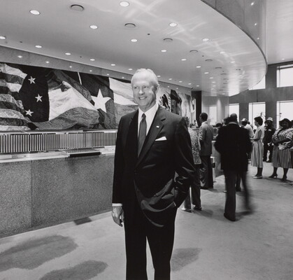 A black-and-white photograph of a White man in a business suit, one hand in his pocket, standing in the lobby of a building with flags adorning the wall in the background.