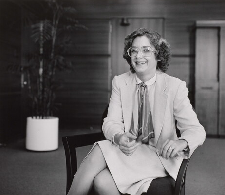 A black-and-white photograph of a seated White woman wearing glasses and a business suit, with a large potted plant in a wood-paneled room.