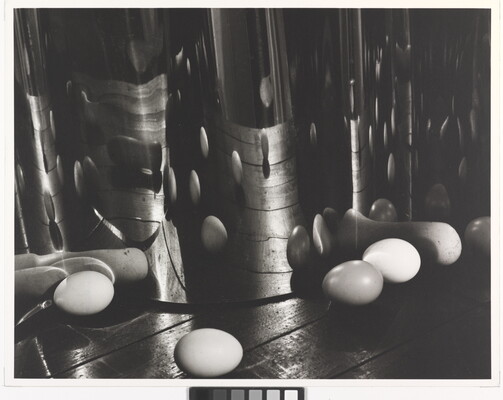 A black-and-white photograph of eggs on a wood surface reflected and distorted by an upright, wavy mirrored surface.