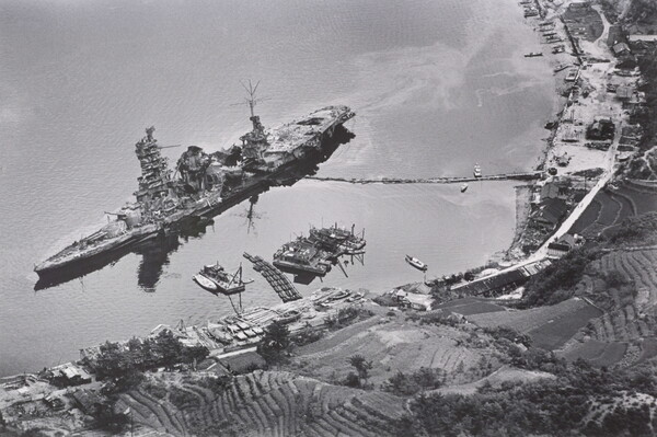 A black-and-white photograph of an aerial view of a large, damaged ship just off a hilly coast.