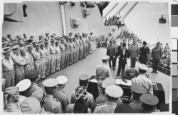 A black-and-white photograph of soldiers standing on the deck of a ship watching a smaller group of figures sign papers at a table.