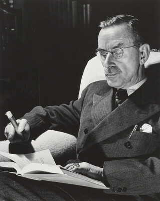 A black-and-white portrait photograph of a seated White man wearing a dark suit and glasses, holding a cigar, and reading a book.