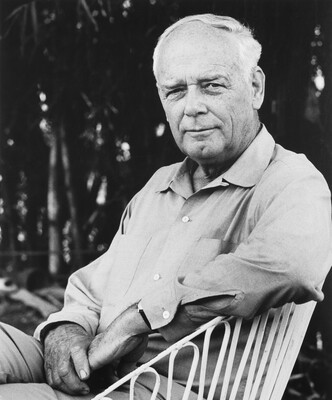 A black-and-white photograph of a seated older, balding White man with a cleft chin wearing a long-sleeved button-down shirt, one arm resting on the back of his chair.