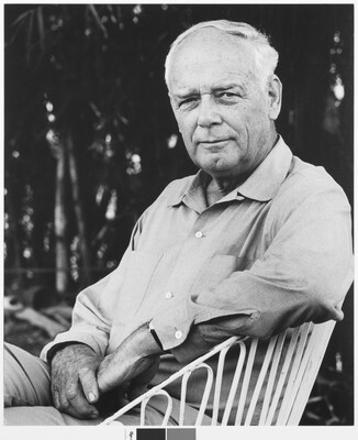 A black-and-white photograph of a seated older, balding White man with a cleft chin wearing a long-sleeved button-down shirt, one arm resting on the back of his chair.