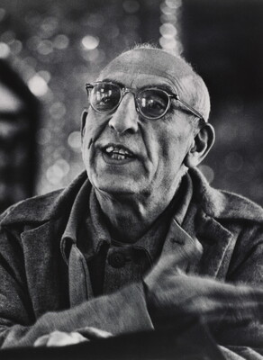 A black-and-white photograph of a balding, medium-skinned man wearing glasses talking and gesturing with his hand.