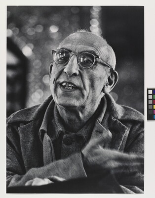 A black-and-white photograph of a balding, medium-skinned man wearing glasses talking and gesturing with his hand.