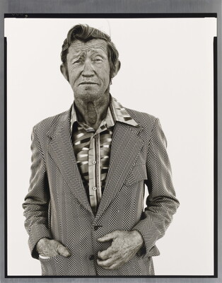 A black-and-white portrait photograph of an older White man with a worn face wearing a patterned, open-collar, button-down shirt under a chevron striped jacket.