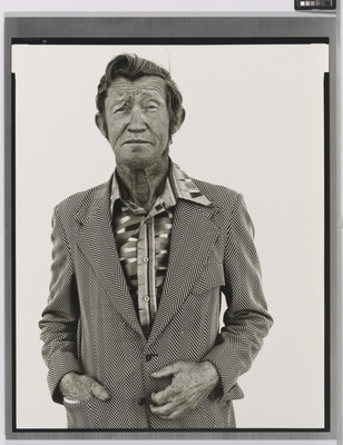 A black-and-white portrait photograph of an older White man with a worn face wearing a patterned, open-collar, button-down shirt under a chevron striped jacket.