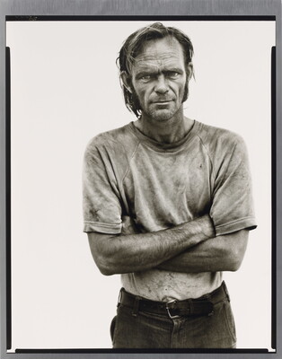 A black-and-white portrait photograph of a White man with unkempt hair in a dirty tee-shirt standing with arms crossed.