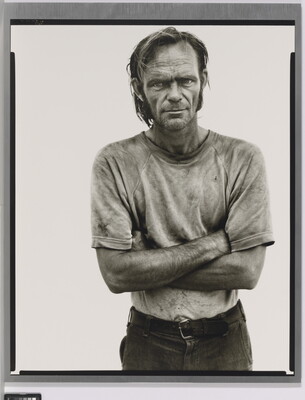 A black-and-white portrait photograph of a White man with unkempt hair in a dirty tee-shirt standing with arms crossed.