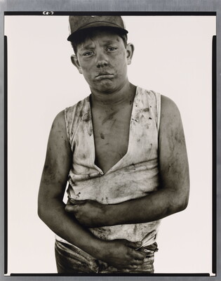 A black-and-white portrait photograph of a dirty young man wearing a baseball hat, a dirty ripped shirt, and holding his arms across his torso.