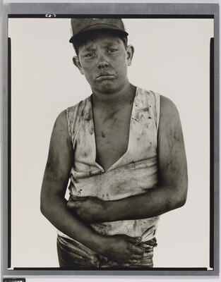 A black-and-white portrait photograph of a dirty young man wearing a baseball hat, a dirty ripped shirt, and holding his arms across his torso.