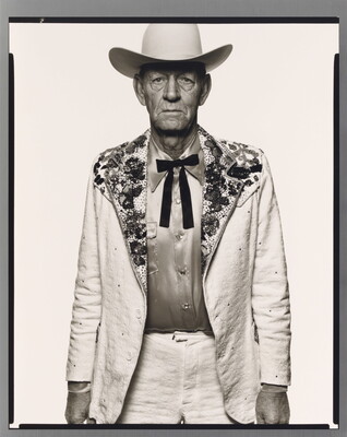A black-and-white portrait photograph of an older White man wearing a white cowboy hat, a white suit jacket with rhinestones on the collar, a satin shirt, and a bow tie.