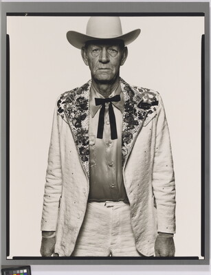 A black-and-white portrait photograph of an older White man wearing a white cowboy hat, a white suit jacket with rhinestones on the collar, a satin shirt, and a bow tie.