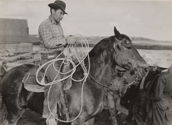 A black-and-white photograph of a light-skinned man in a brimmed hat holding a lasso on horseback.