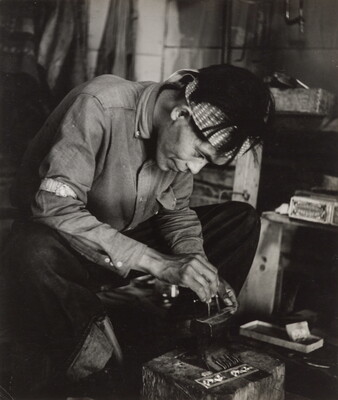 A black-and-white photograph of a seated Native American man bent over an anvil in a workshop.