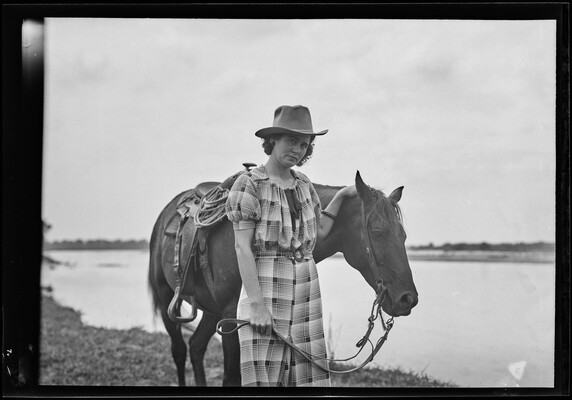 A black-and-white image of a White woman wearing a cowboy hat and plaid dress holding the reins of the horse standing next to her.