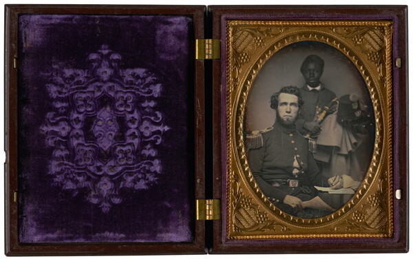 A black-and-white photograph in a hinged case with an oval frame of a seated White man in a military uniform and a  young Black boy standing behind him.